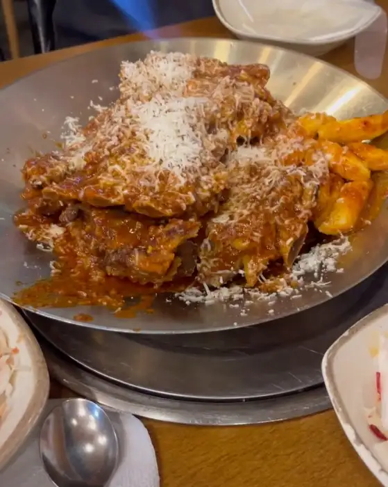철판 위에 뼈찜이 담겨 있고, 그 위로 치즈 가루가 뿌려져 있습니다. 떡도 함께 보입니다.