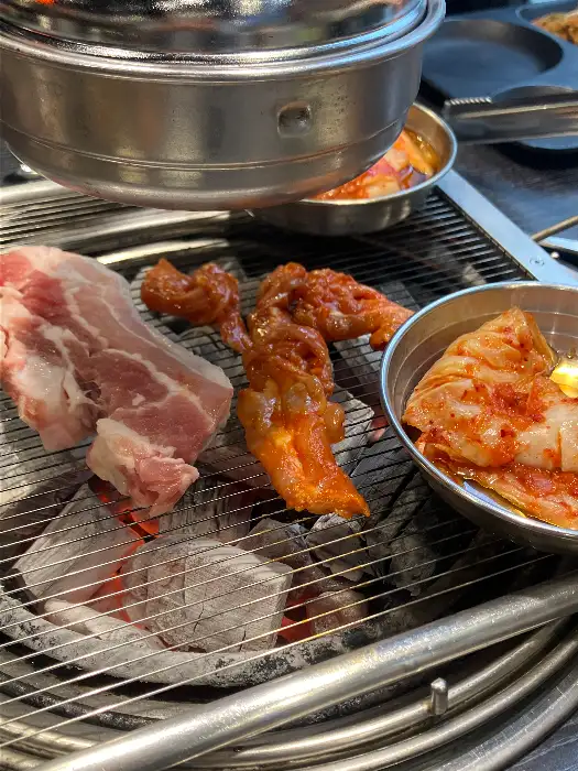 불판 위에 놓인 삼겹살, 양념된 닭고기, 김치, 버섯, 마늘
