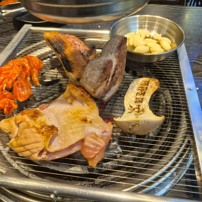 불판 위에 고기, 버섯, 마늘, 양념된 닭고기가 익고 있는 모습