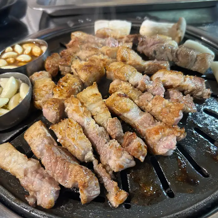 잘 구워진 돼지고기가 숯불 위에서 노릇노릇하게 익어가고 있다. 기름이 떨어지면서 숯불과 만나 연기가 피어오르는 모습이 보인다.