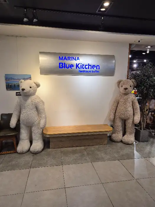 MARINA BLUE KITCHEN이라고 쓰인 간판이 걸려 있고, 양쪽에 큰 곰 인형이 서 있는 입구.