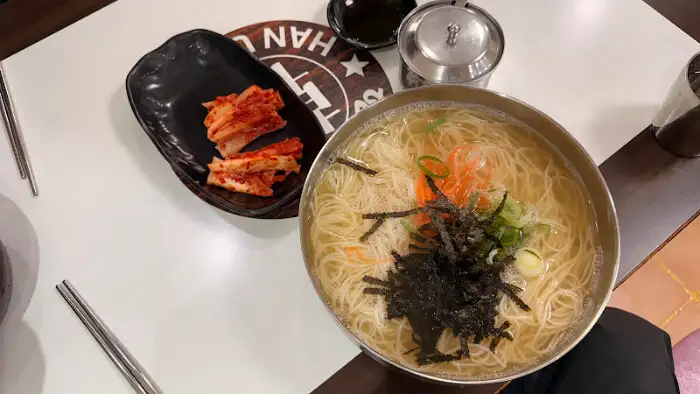 테이블 위에 놓인 잔치국수와 김치. 놋그릇에 담긴 국수와 검은색 접시에 담긴 김치가 대비를 이룬다.