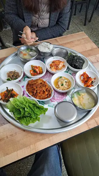 식탁 위에 쟁반에 여러 반찬들이 놓여있고, 한쪽에는 신선한 채소가 담겨 있다.
