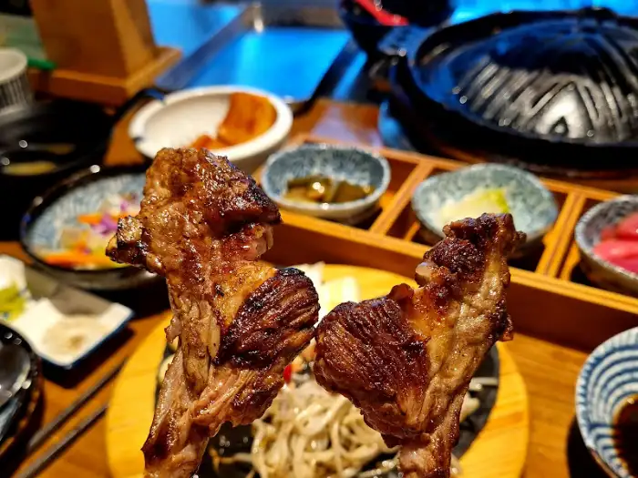 손가락으로 잡은 양갈비 두 덩어리가 클로즈업되어 있다. 배경으로는 반찬과 불판이 흐릿하게 보인다.