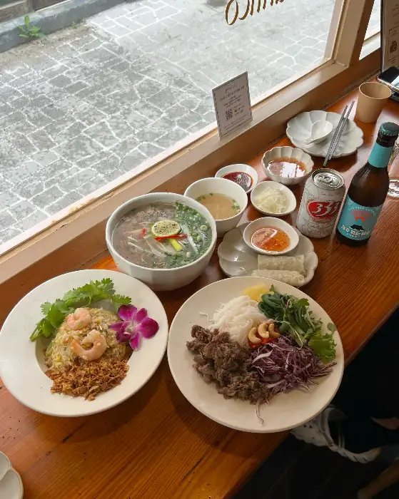 창가 테이블에 놓인 쌀국수, 볶음밥, 샐러드, 그리고 맥주와 스프링롤 등이 푸짐하게 차려진 모습