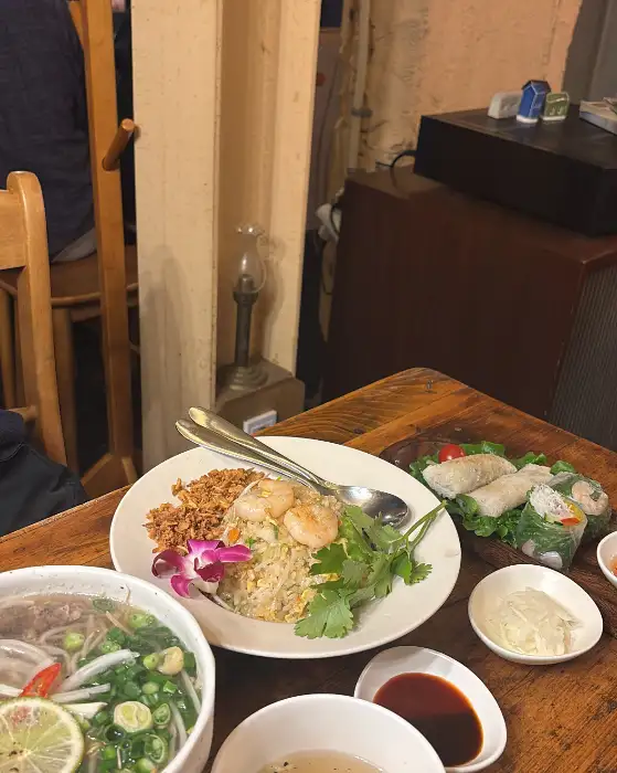 테이블 위에는 쌀국수, 볶음밥, 스프링롤 등이 놓여 있고, 한쪽에는 붉은색 꽃 장식이 있는 모습