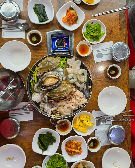 해물탕 냄비를 중심으로 여러 가지 반찬 그릇들이 놓여 있습니다. 전복, 새우, 조개 등 신선한 해물이 가득 담긴 해물탕이 중심에 있습니다.