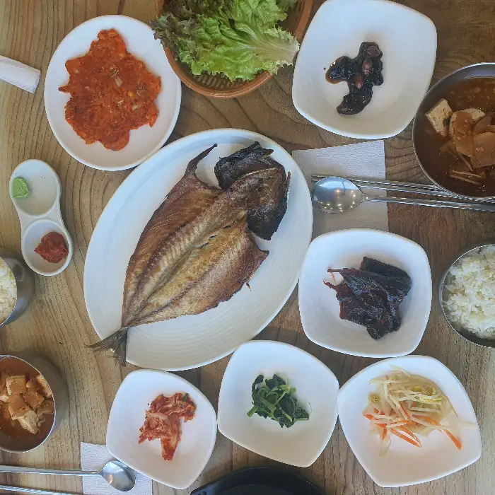 식탁 위에 차려진 푸짐한 한상차림. 노릇하게 구워진 고등어와 여러 가지 반찬들이 정갈하게 담겨 있다.