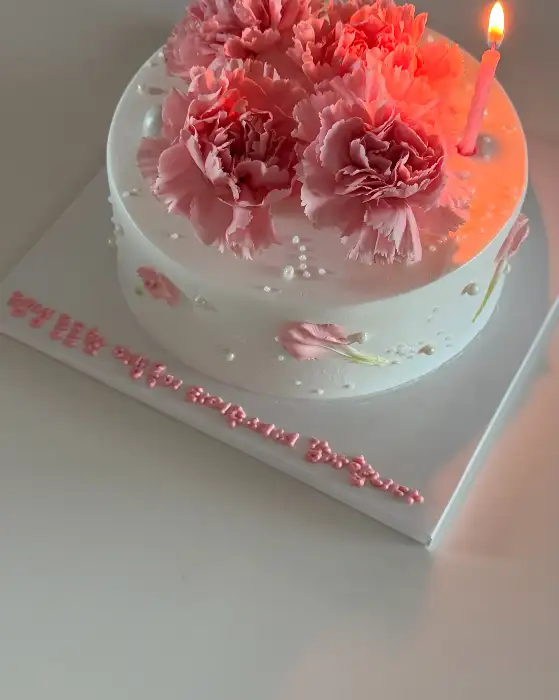 핑크색 카네이션으로 장식된 하얀색 케이크 위에 핑크색 초가 꽂혀있고, 케이크 옆면에는 핑크색 레터링이 새겨져 있는 모습