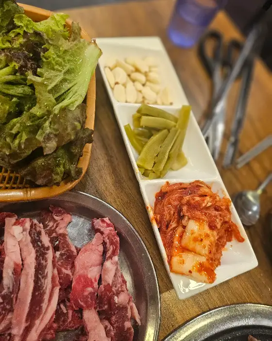 신선한 채소와 쌈무, 김치, 마늘 슬라이스가 정갈하게 담긴 접시와 신선한 생고기가 준비된 모습