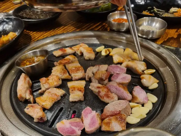잘 익은 다양한 부위의 돼지고기 조각들이 불판 위에 가지런히 놓여 있고, 곁들여 먹을 마늘과 소스도 보입니다.