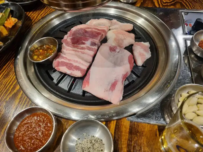 두툼한 생 돼지고기 덩어리가 불판 중앙에 놓여 있고, 주변에는 소스와 마늘이 준비되어 있습니다.