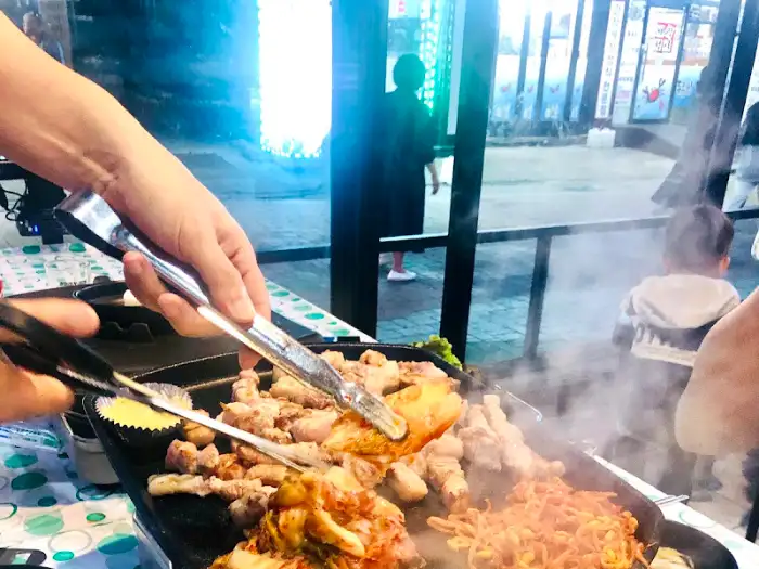 집게로 삼겹살을 뒤집고 있는 모습. 테이블에는 볶음김치와 콩나물이 보인다.