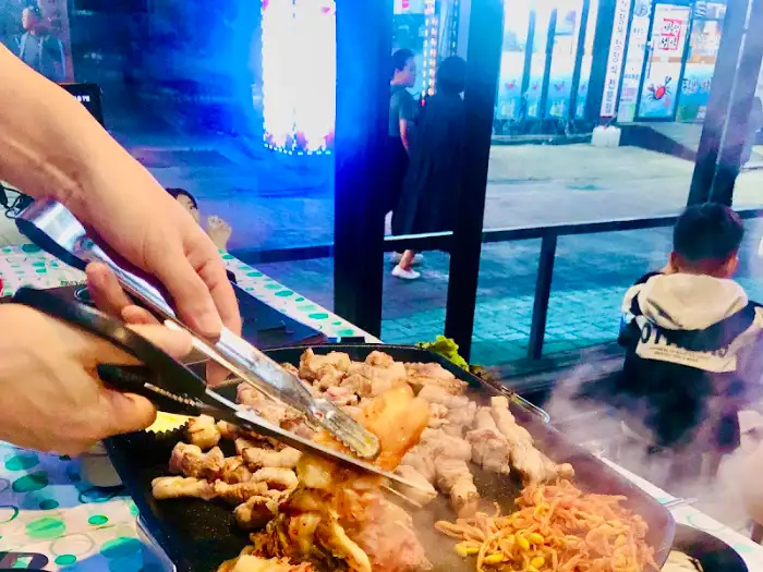 집게로 삼겹살 조각을 들어 올리는 손. 불판에는 콩나물과 김치가 익어가고 있다.