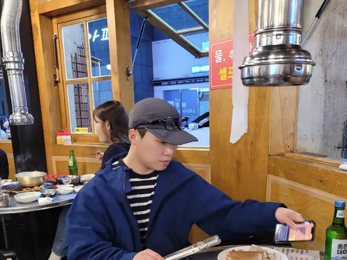 테이블에 앉아있는 사람들 모습, 고기 불판과 술병이 보인다.