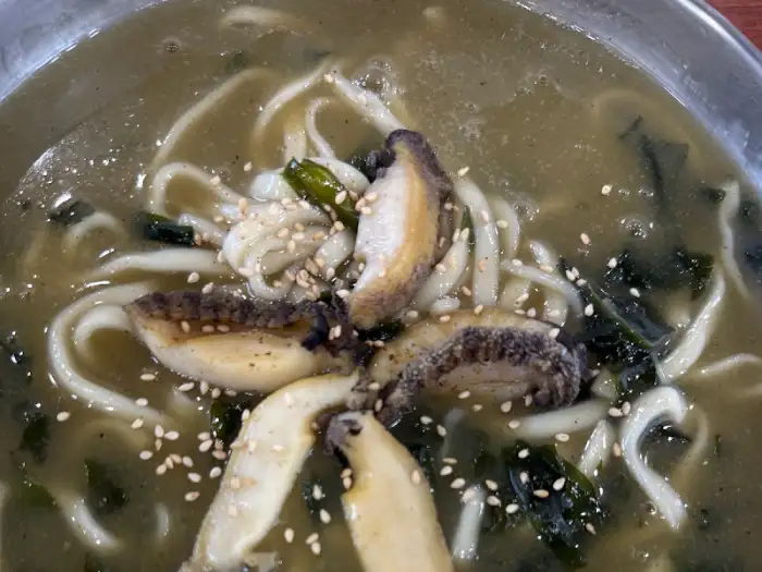 전복과 해물이 들어간 칼국수 근접 촬영 샷, 면발과 국물, 해물이 조화롭게 보임.
