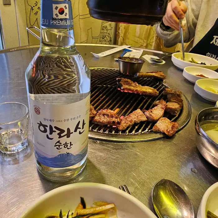 테이블 위에 놓인 한라산 소주 병과 잔, 그리고 고기가 구워지고 있는 불판