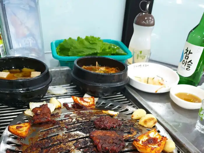 테이블 위에 차려진 푸짐한 한상차림. 연탄 불판 위에 고기와 채소가 올라가 있고, 곁들임 음식과 소주병이 보인다.