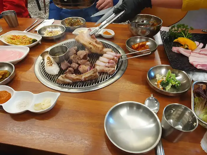 직원분이 집게를 이용해 숯불 위 고기를 능숙하게 뒤집고 있습니다. 테이블 위에는 다양한 밑반찬과 소스 그릇들이 보입니다.