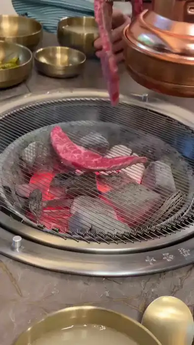 집게로 두툼한 소고기 한 점을 들어 숯불 위로 옮기고 있는 모습. 붉은 숯불이 활활 타오르고 있다.