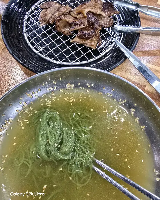 금빛으로 빛나는 숯불고기 접시와 젓가락으로 집어 올린 푸른 빛의 냉면 면발
