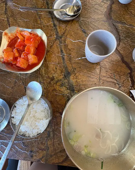 새빨간 깍두기와 함께 놓인 설렁탕과 밥 한 공기, 그리고 놋수저와 컵이 정갈하게 차려진 모습