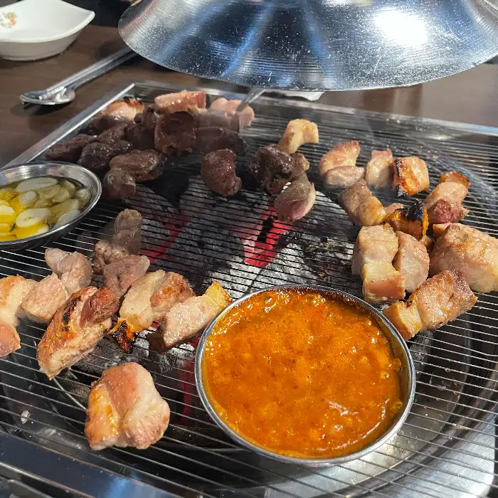 다양한 부위의 흑돼지가 숯불 위에서 구워지고 있는 모습, 멜젓 소스도 함께 보인다