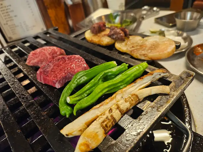 불판 위에 큼직한 소고기 덩어리와 길쭉한 청양고추, 통으로 구워지는 새송이버섯이 놓여 있습니다.