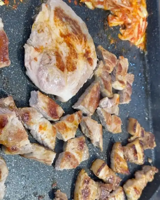 불판 위에 두툼한 목살 한 점과 잘게 썰어진 삼겹살 조각들이 올라가 있습니다. 옆에는 볶은 김치와 콩나물이 보입니다.