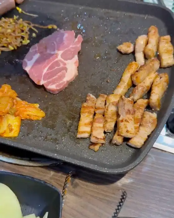 넓은 불판 위에 다양한 고기 조각과 김치, 콩나물 무침이 올려져 있습니다. 고기들은 노릇하게 익어가고 있습니다.