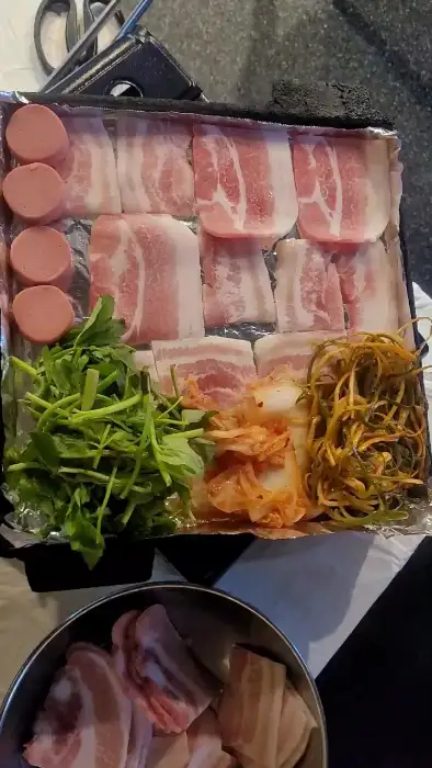불판 위에 놓인 삼겹살, 김치, 미나리, 소시지