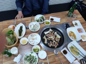현대식육식당 테이블 위 가스 철판에 구워지고 있는 삼겹살과 쌈 채소, 밑반찬들
