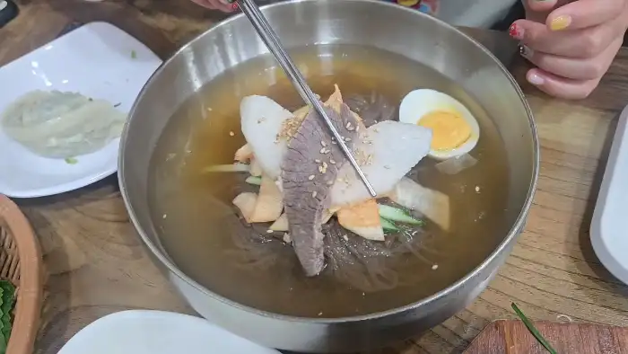놋그릇에 담긴 평양냉면의 면발과 고명을 집게로 건져 올리는 모습