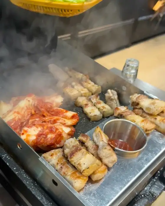 금돈집 광명하안점에서 불판 위에 삼겹살, 김치, 미나리가 함께 구워지고 있는 모습