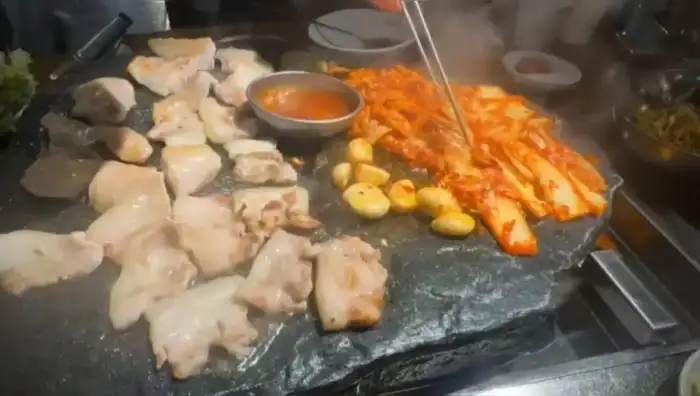 석암생소금구이 진주하대점의 뜨거운 돌판 위 고기, 김치, 마늘 굽는 모습